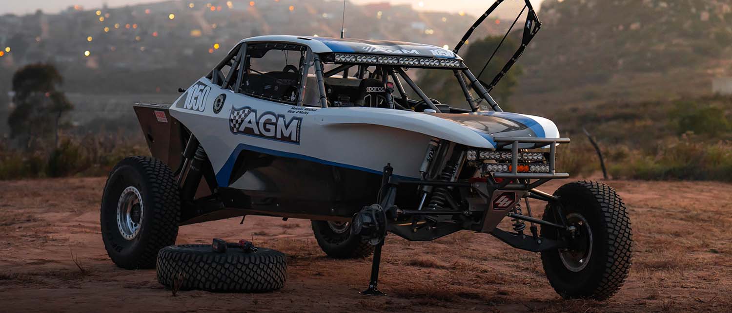 A white and blue off-road racing buggy with 'AGM' branding, parked on a dirt track during sunset with its spare tire on the ground and being lifted by an AGM Manual Jack Pro. 