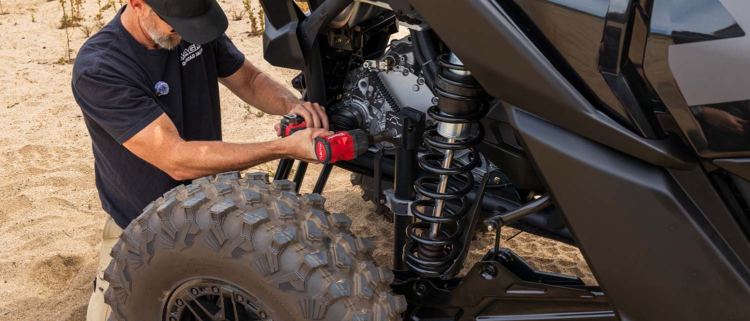 A man in a black t-shirt and cap uses a red cordless impact wrench to operate an AGM Manual Jack Sport lifting a black off-road UTV vehicle on a sandy surface.