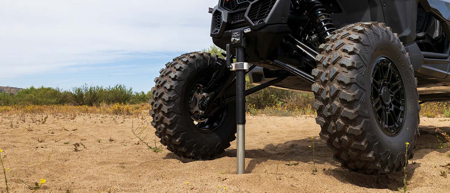 An AGM Manual Jack sport lifting the entire front end of a black Can-Am X3 off-road vehicle with large knobby tires parked outdoors on sandy ground.
