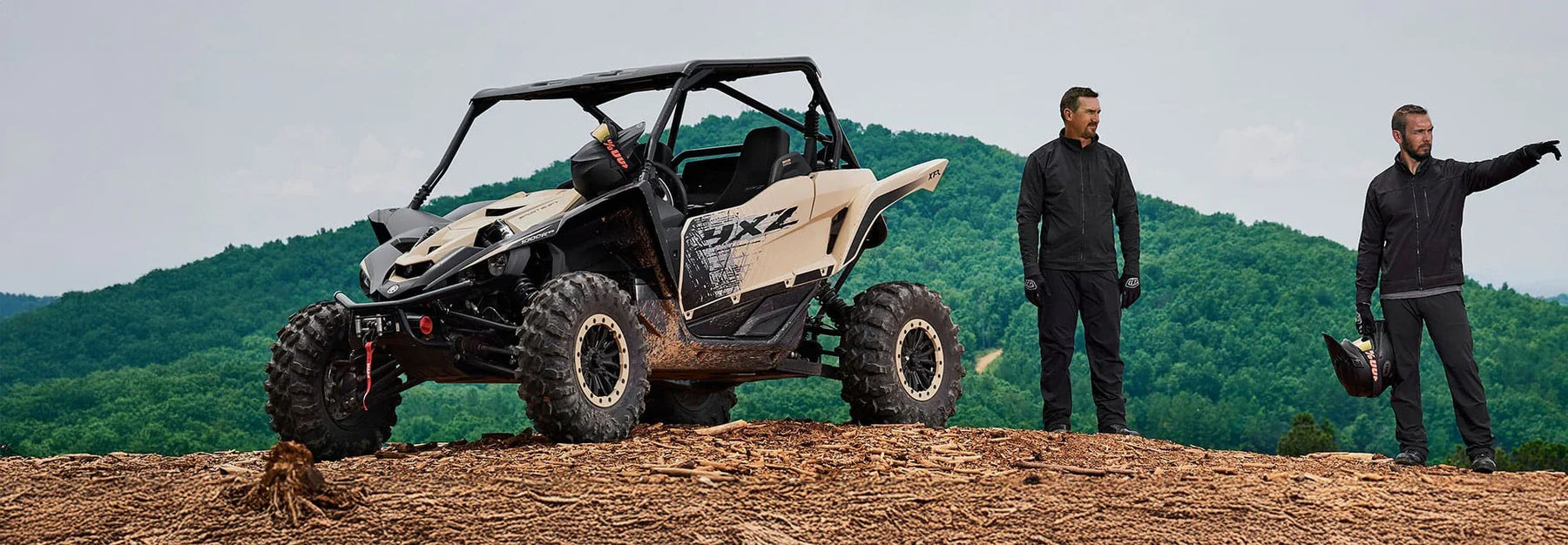 Two people standing next to a large off-road vehicle on a dirt hill with green mountains in the background.