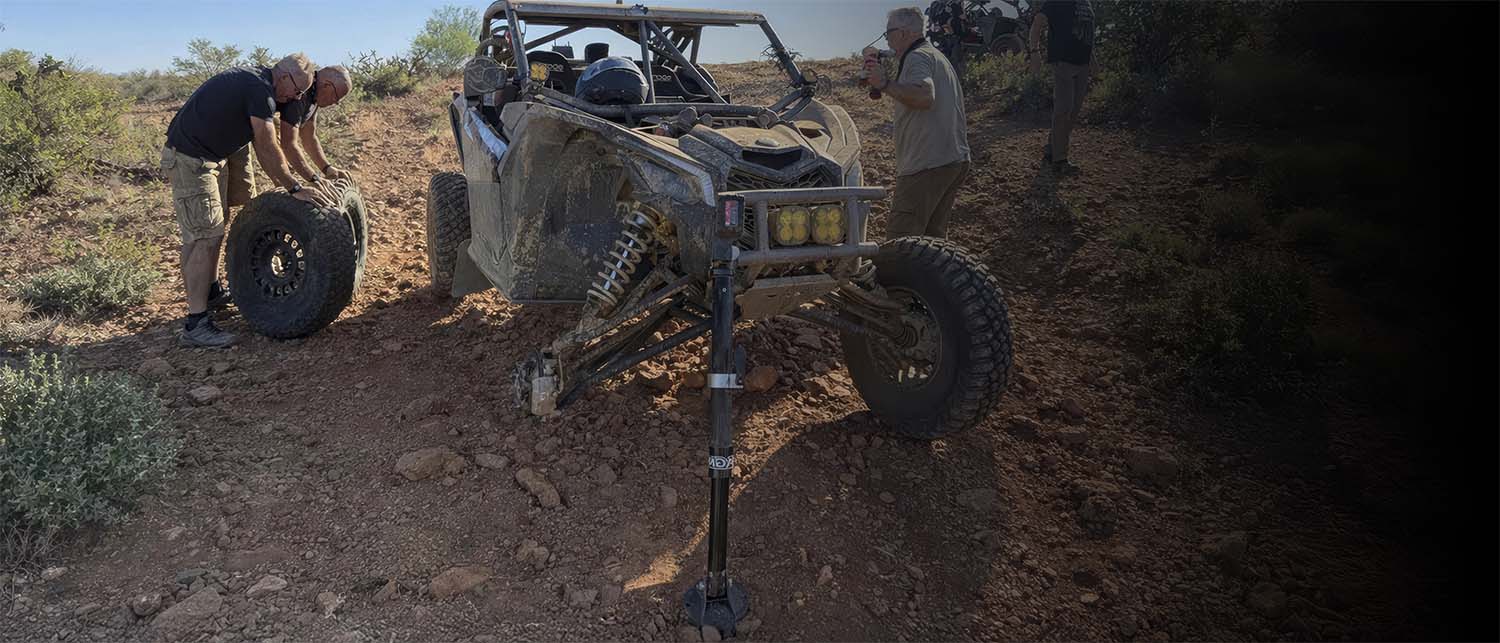 A rugged off-road UTV being held up by an AGM Electric Jack. Parked on a dirt trail, with two people handling a spare tire in a desert setting.