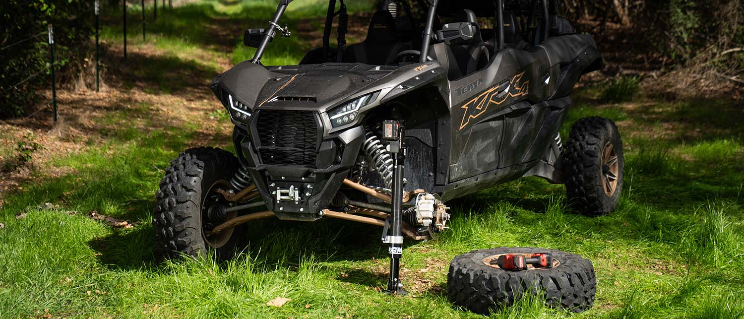 A black and gray Kawasaki KRX with the front suspension lifted by an AGM Electric jack. Next to a spare tire, parked on grass in a wooded outdoor setting.