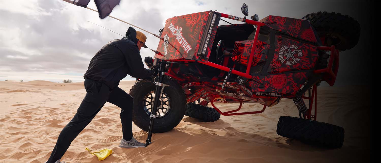 A red and black off-road desert buggy with large knobby tires is shown in a sandy desert under an overcast sky, with a person using an AGM Electric Jack to roll it back onto its wheels. 