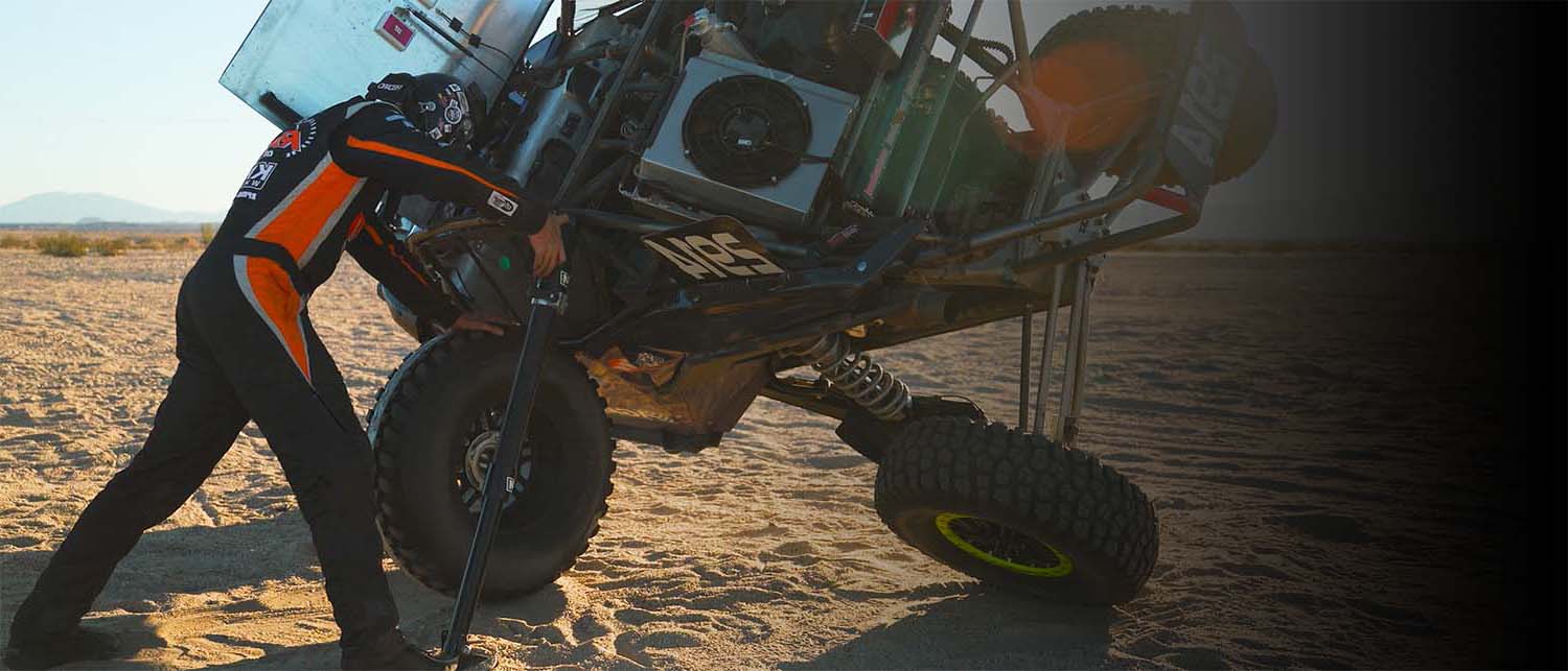 A person in black and orange clothing uses an AGM Electric Jack to roll a vehicle back onto its wheels in a sandy desert setting.