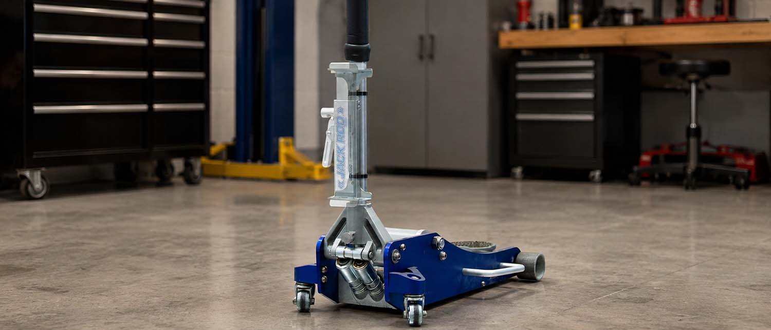 A blue and silver floor jack with an AGM Jack Rod mount zip tied to the handle, positioned on a concrete workshop floor with garage tools and cabinets in the background.