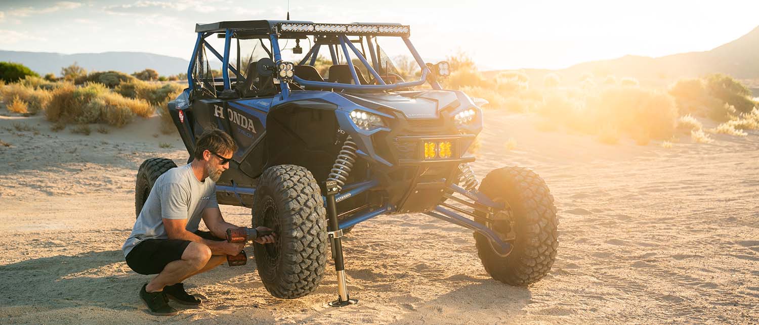 A man crouches beside a blue honda off-road side-by-side vehicle in a sandy desert setting during golden hour, with the front tire lifted on an AGM Manual jack sport.