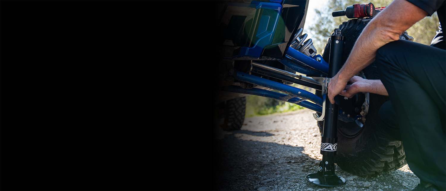 A person crouches beside a blue and black off-road vehicle on a dirt trail, preparing an AGM Manual Jack Pro to lift the vehicle's front end.