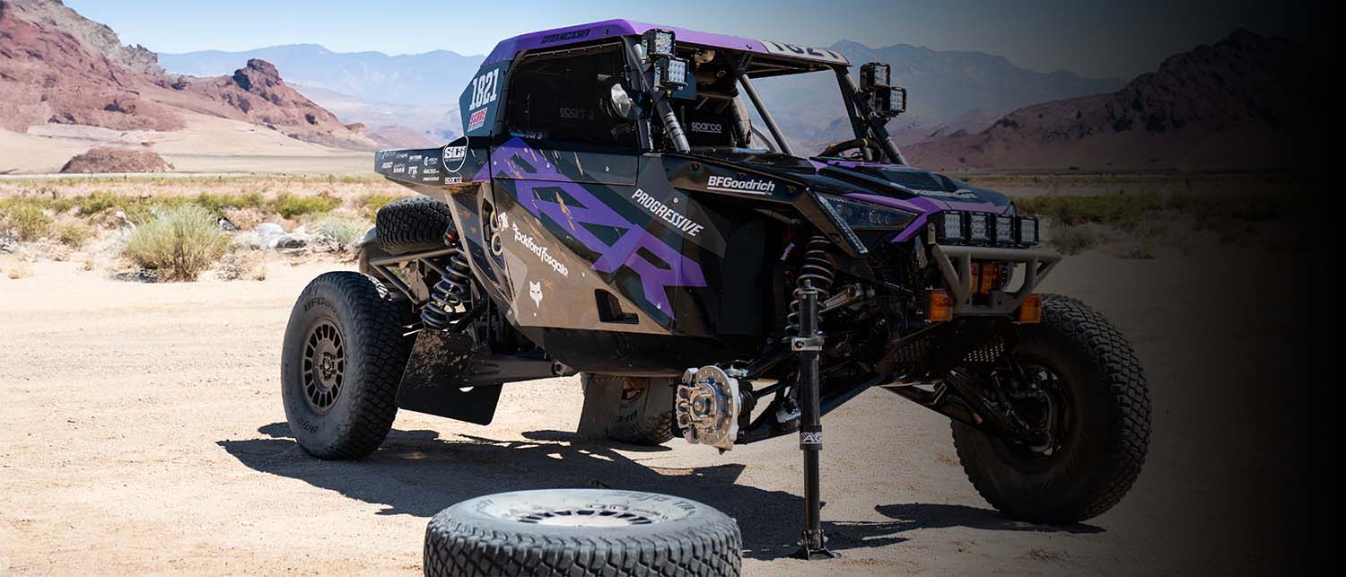 A black and purple Factory Polaris RZR race car with large off-road tires is parked on a sandy desert road with mountains in the background. A spare tire is resting in the foreground. Car is being held up by an AGM Manual Jack pro