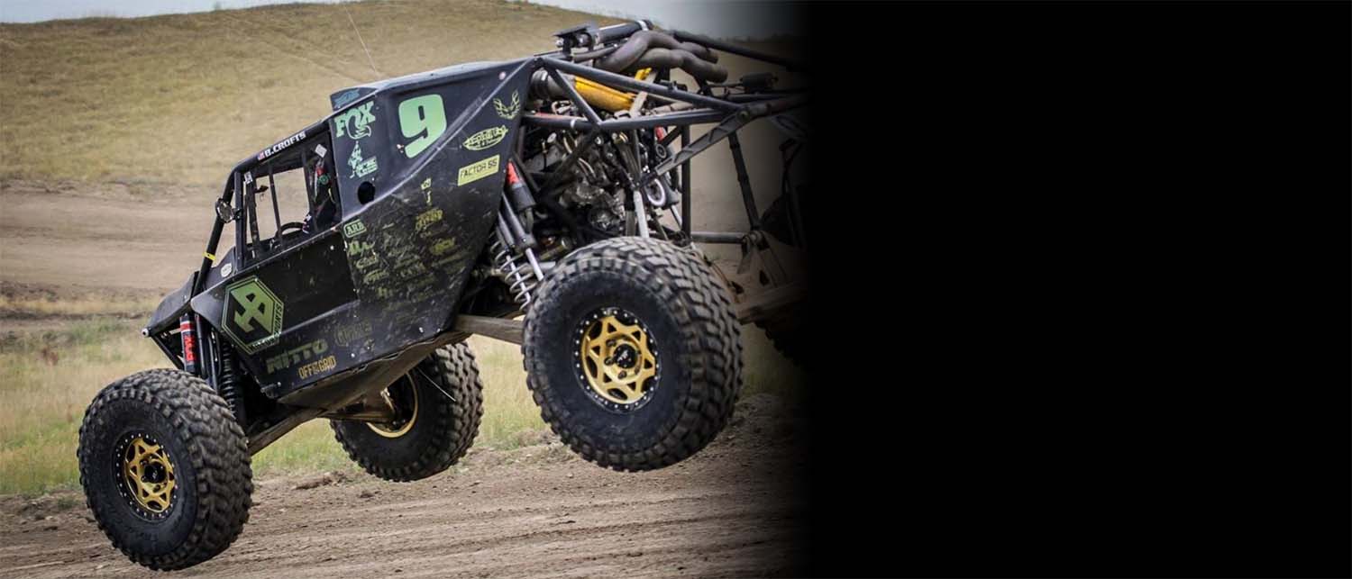 A black off-road racing buggy with large off-road tires, gold wheels and an AGM Manual Jack Pro mounted on the back, captured mid-air on a dirt track.