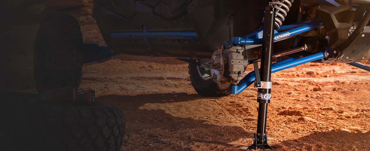 A close-up outdoor shot of an off-road vehicle's blue suspension components, including a shock absorber and control arm, being supported in the air by an AGM Manual jack pro against a sandy, dusty ground.