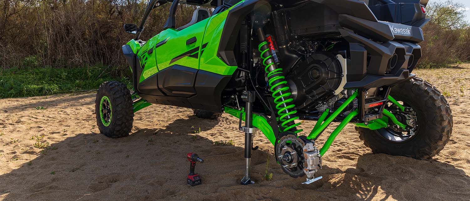 A bright green and black Kawasaki side-by-side UTV parked outdoors on sandy ground with its rear wheel off the ground, supported by an AGM Manual jack sport. A cordless drill lies on the ground nearby, and dry brush and trees are visible in the background.