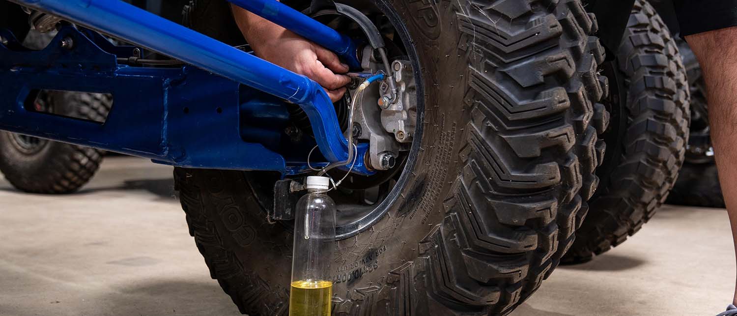A close-up of a person's hand working on a vehicle's wheel hub and brake assembly, with a bottle of yellow oil placed on the ground below the tire.