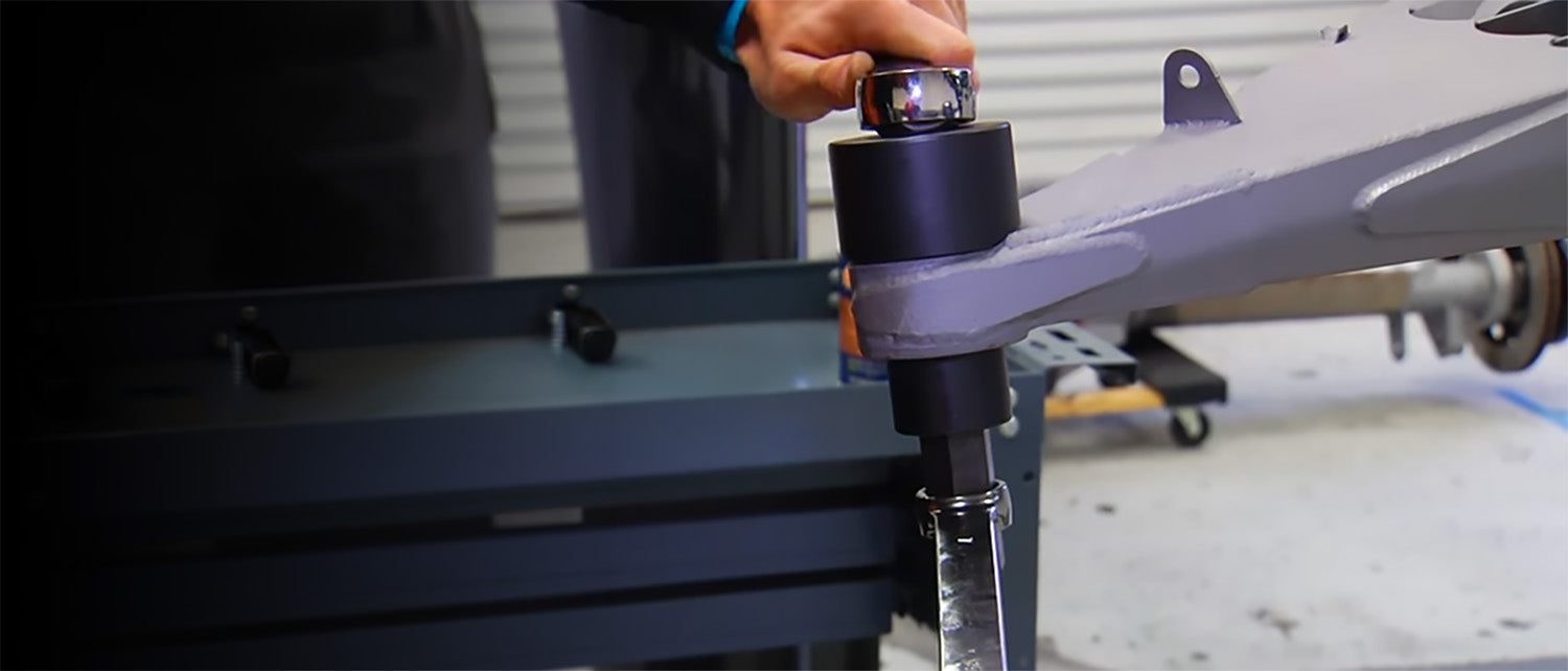 A close-up shot of an AGM Uni-Ball tool on a gray metal lower control arm, with a hand partially visible at the top in an race workshop setting.